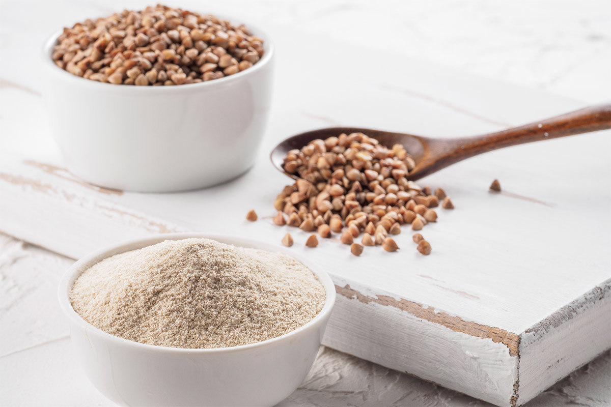 Buckwheat grain and buckwheat flour are in two white bowls. Some grains are scattered on a white board next to the bowls and wooden spoon | Girl Meets Food