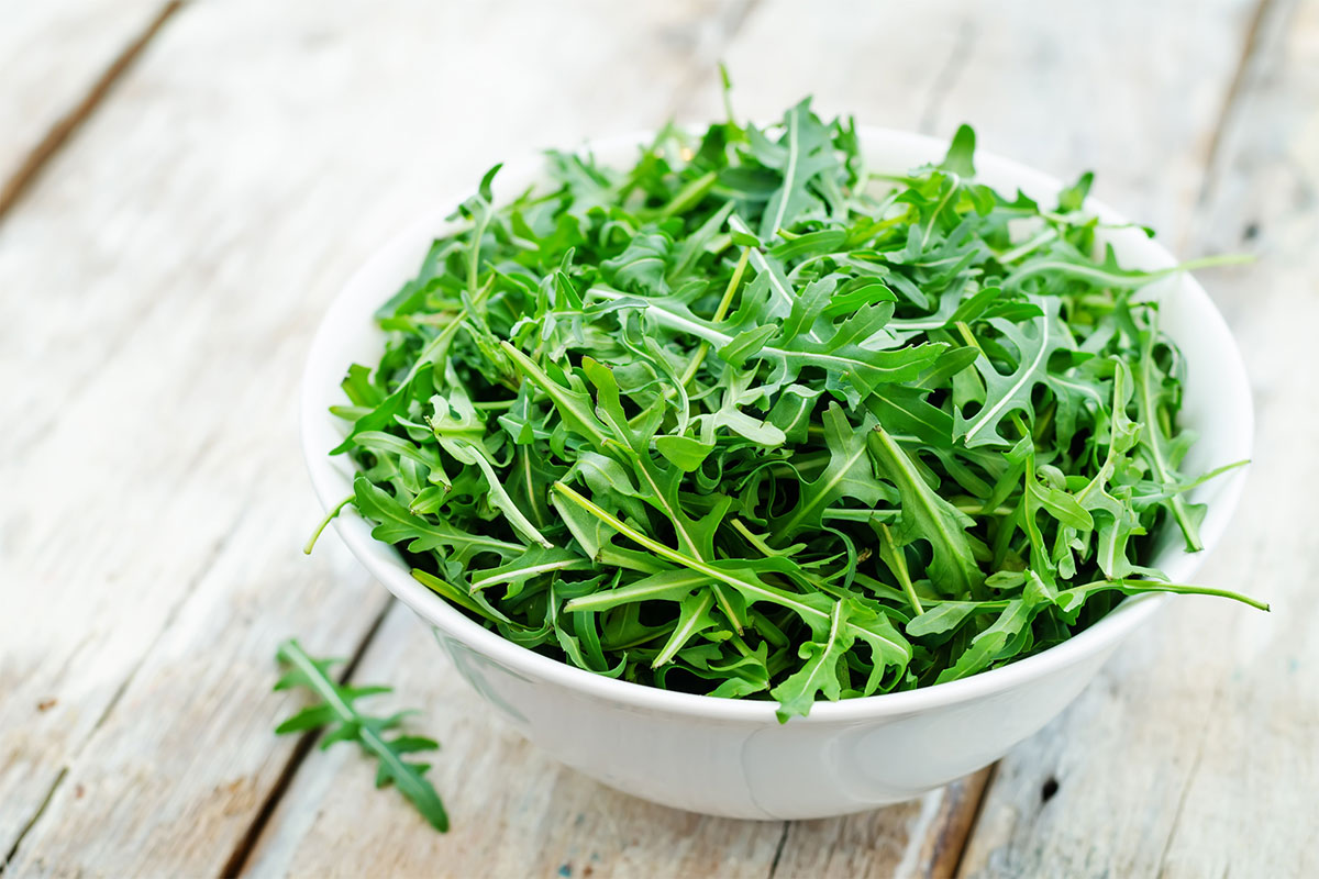 Fresh arugula leaves are in a white bowl that is standing on a wooden surface | Girl Meets Food