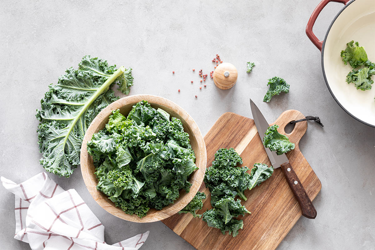 Fresh kale leaves are in a wooden bowl and on a chopping board with a knife and towel | Girl Meets Food