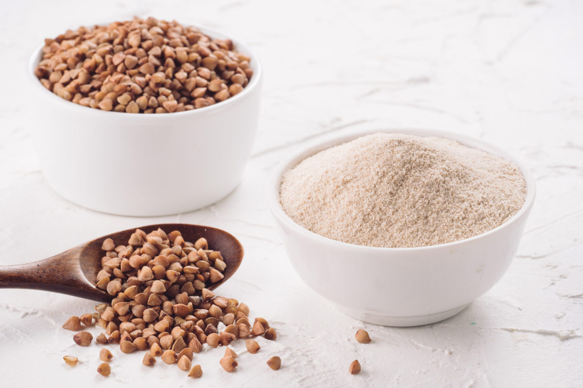 Buckwheat grain and buckwheat flour are in two white bowls. Some grains are scattered on a white surface next to the bowls and wooden spoon | Girl Meets Food
