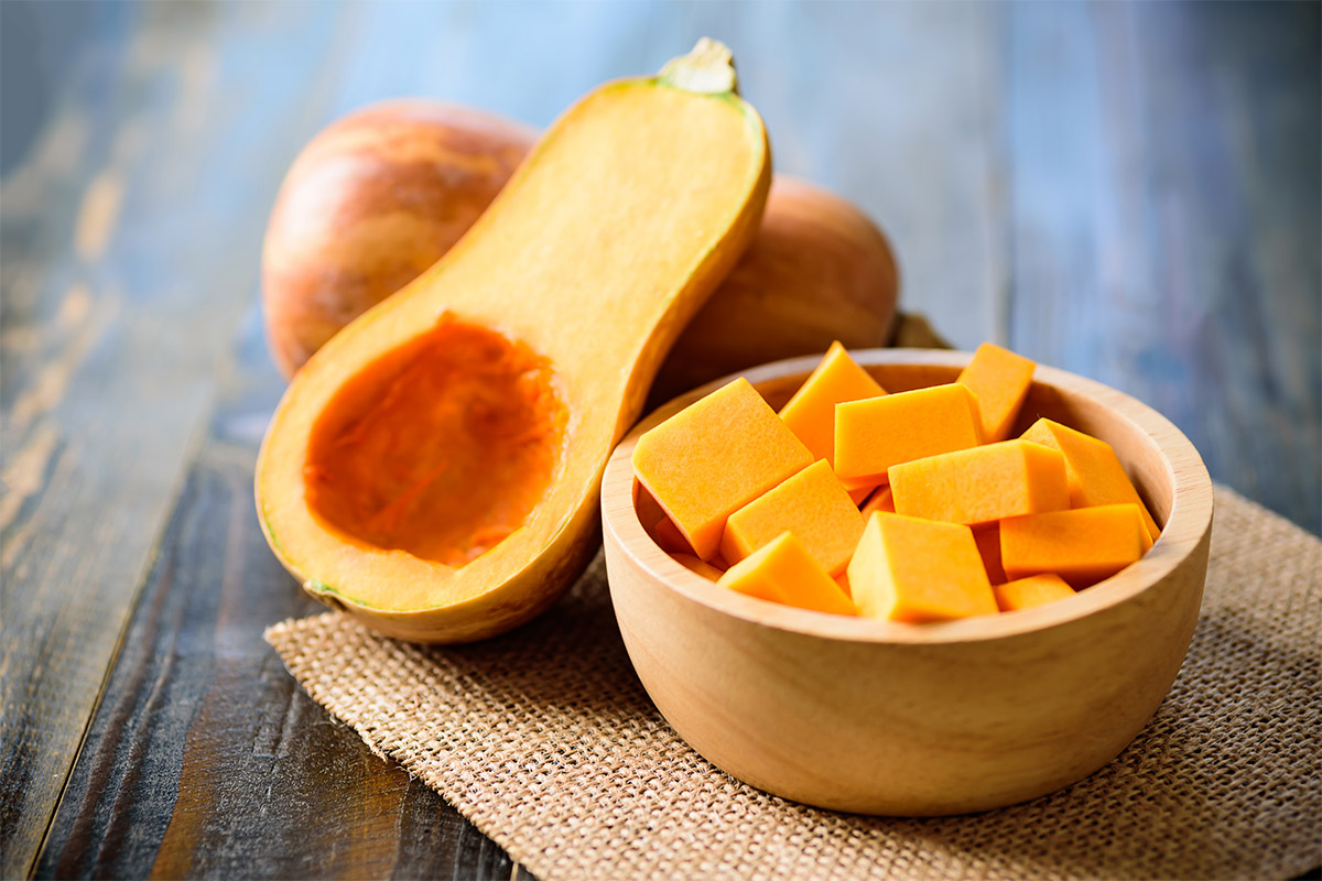 A wooden bowl of butternut squash cubes stands on a linen piece of cloth. There are a butternut squash and a half next to it | Girl Meets Food
