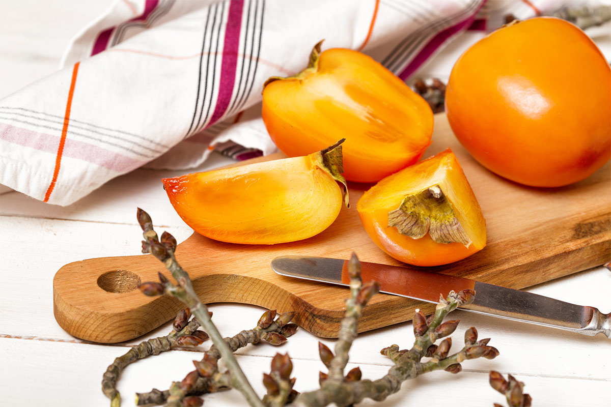 Chopped and a whole persimmon are on a cutting board that stands on a white surface. There is a towel next to it | Girl Meets Food