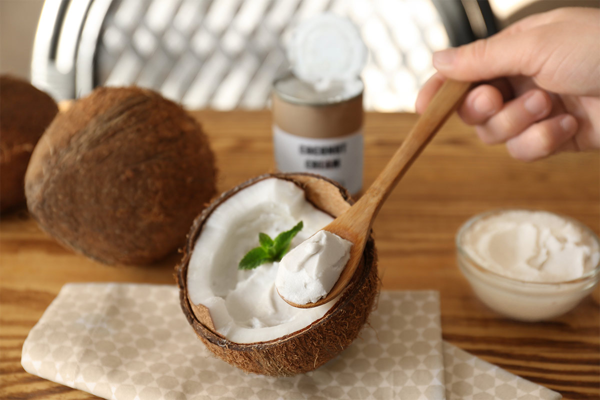 Hand holding a wooden spoon of coconut cream. In the background are some coconuts, a jar and a bowl of coconut cream | Girl Meets Food