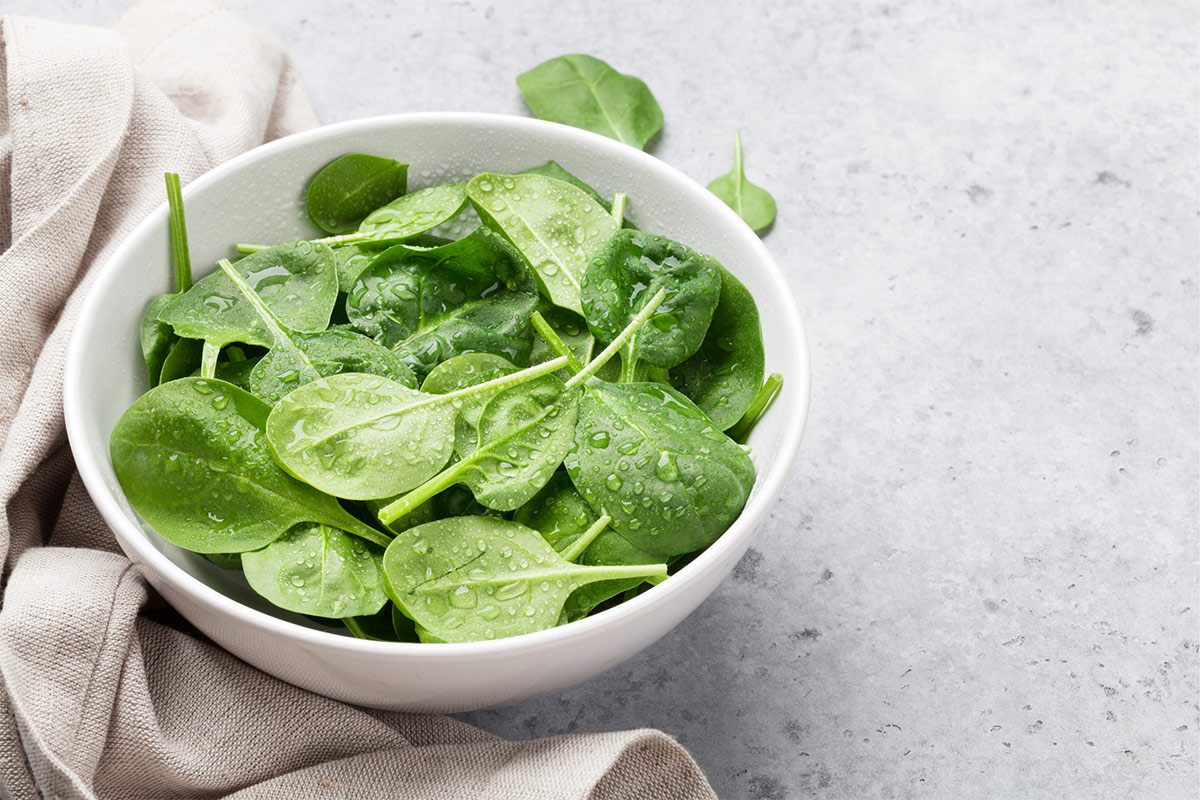 A white bowl of fresh spinach stands on grey fabric and murble surface | Girl Meets Food