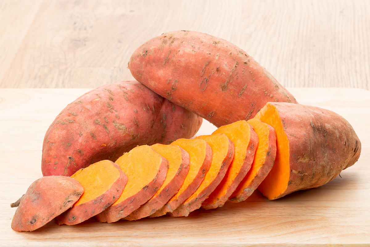 Two raw sweet potatoes and one sliced potato lie on a wooden surface | Girl Meets Food