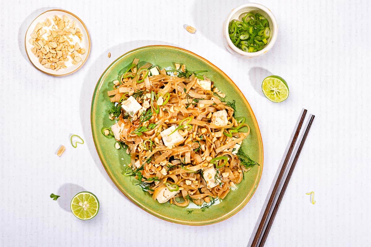 A green oval plate of vegan pad thai, black chopsticks and some other ingredients lie on a white surface | Girl Meets Food