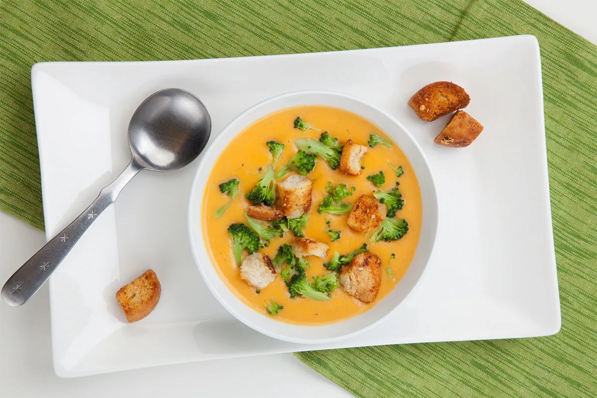 A bowl of broccoli cheese soup, a tablespoon and a few croutons stand on a white plate that is on the table | Girl Meets Food