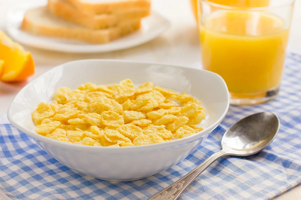 A bowl of corn flakes with milk is on a checkered tablecloth. There is a tablespoon, a glass of orange juice, a plate with toasts and some orange wedges around the bowl | Girl Meets Food