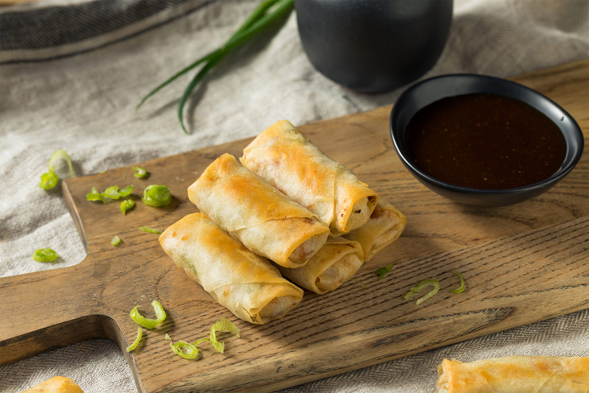 Five egg rolls and a small black bowl with soy sauce are on a cutting board that is on a tablecloth | Girl Meets Food