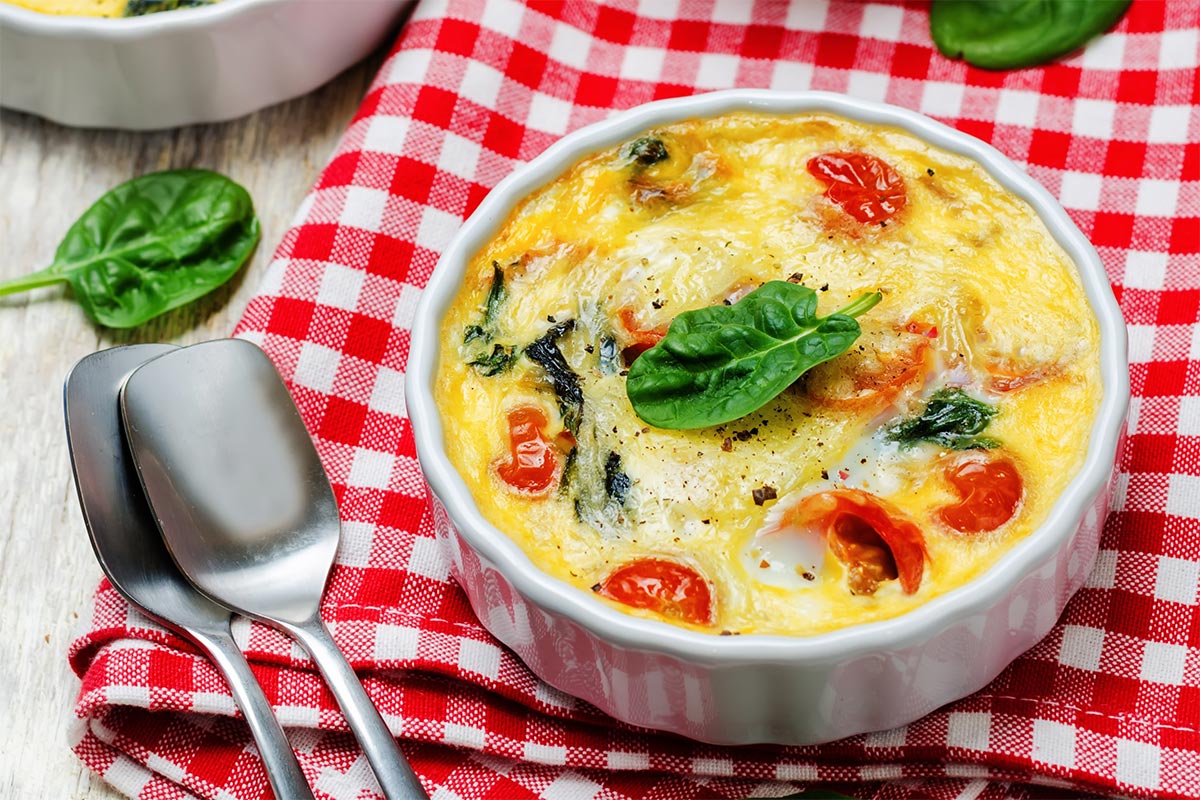 A round baking dish with frittata is on a checkered tablecloth. There are two tablespoons and some spinach leaves next to it | Girl Meets Food