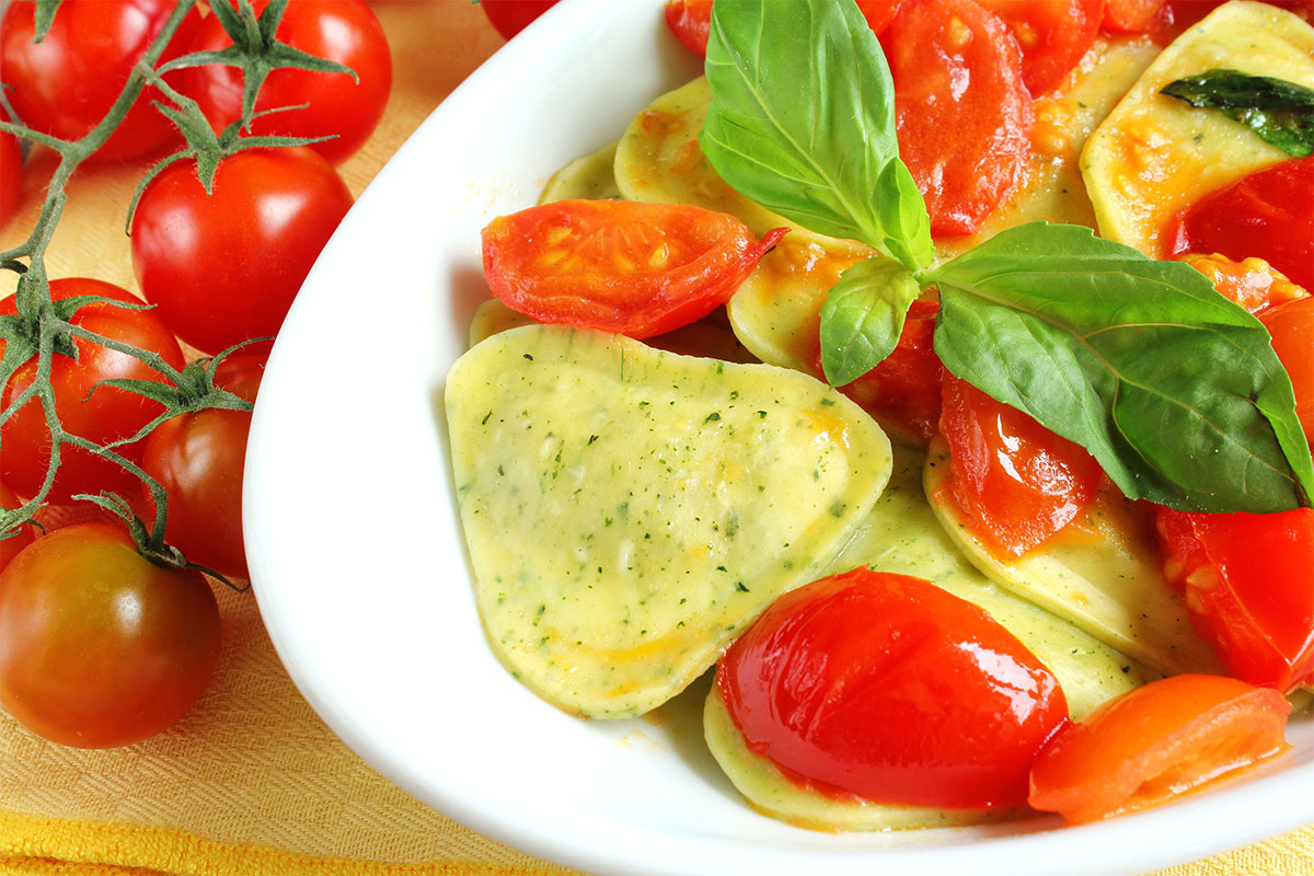 A white plate of ravioli topped with tomato wedges and basil. There are other tomatoes alongside the plate | Girl Meets Food