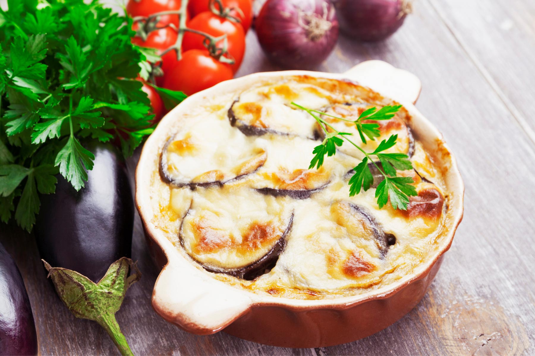 A round red dish with moussaka is on a wooden surface. Alongside it there are red onions tomatoes, cilantro and eggplant | Girl Meets Food