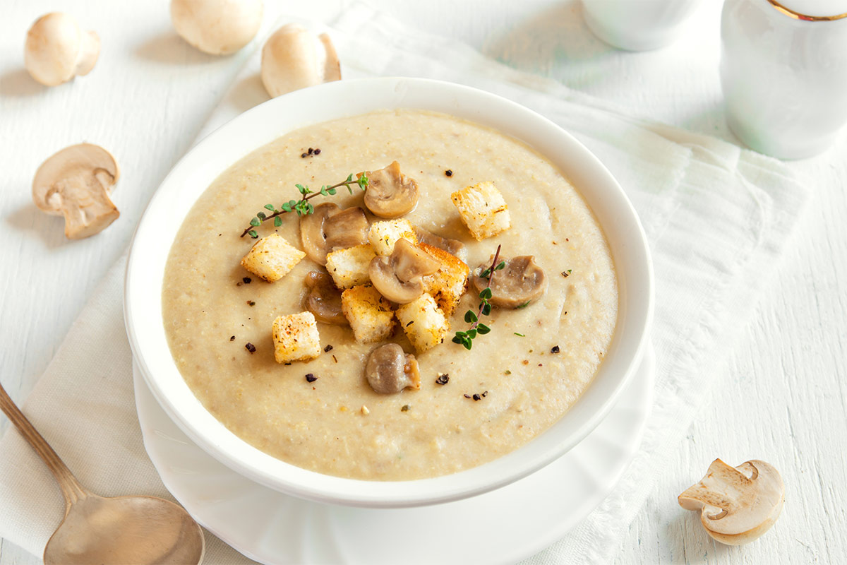 A bowl of mushroom cream soup stands on a white plate that is on a table. There is a tablespoon and some scattered mushrooms around the bowl | Girl Meets Food