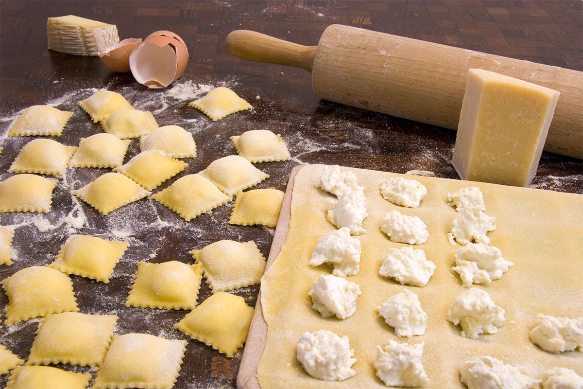 On a black surface there are sculpted ravioli, a board with raw ravioli, rolling pin and cheese | Girl Meets Food