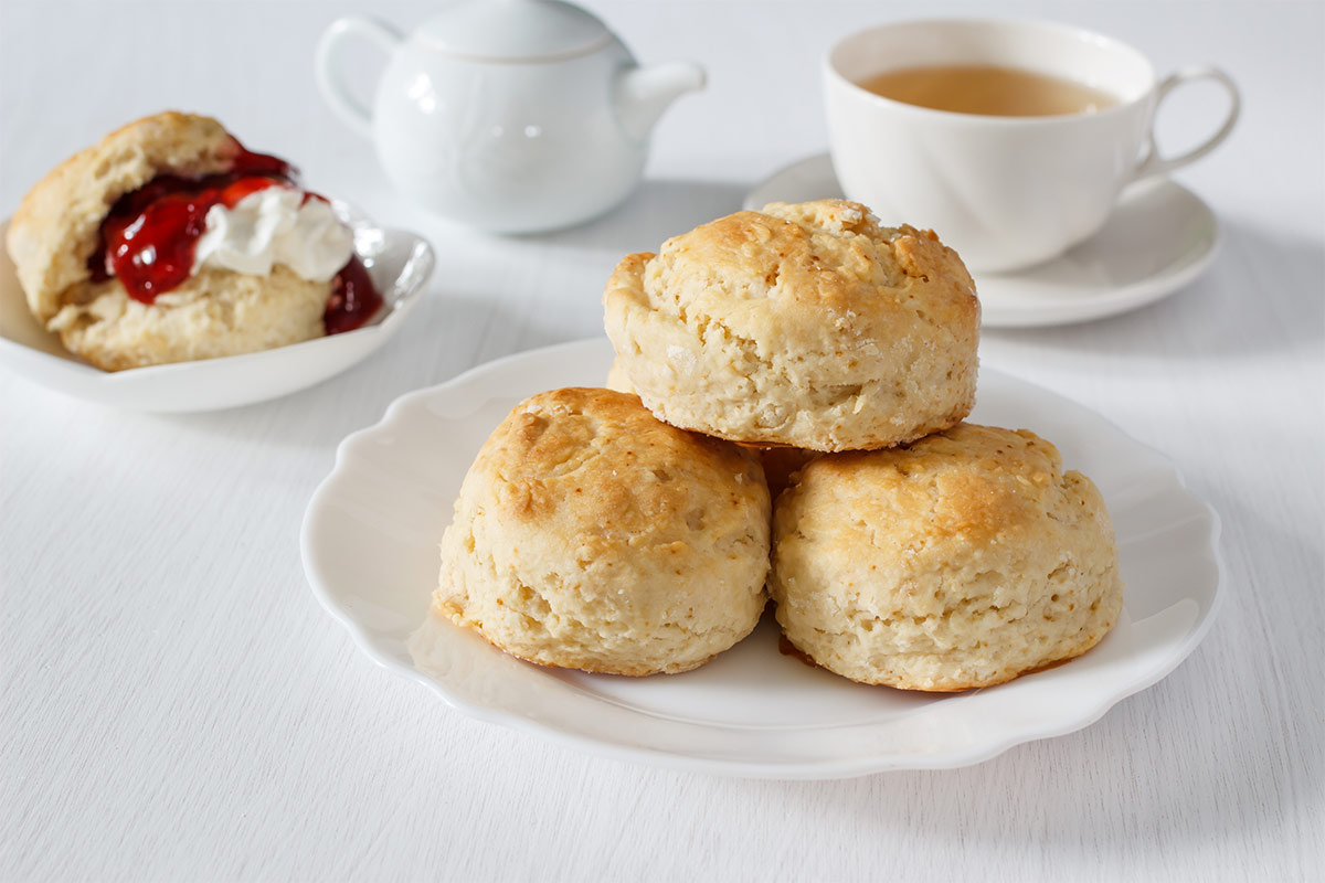 A plate with scones, a plate with scone with topping, a teapot  and a cup of tea are on a white tablecloth | Girl Meets Food