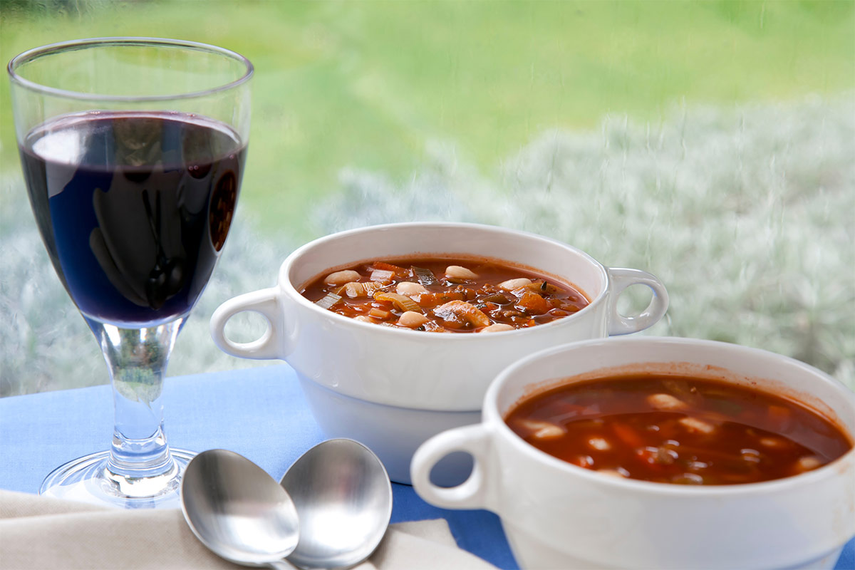 Two bowls of minestrone soup, two tablespoons and a glass of red wine are on a table | Girl Meets Food