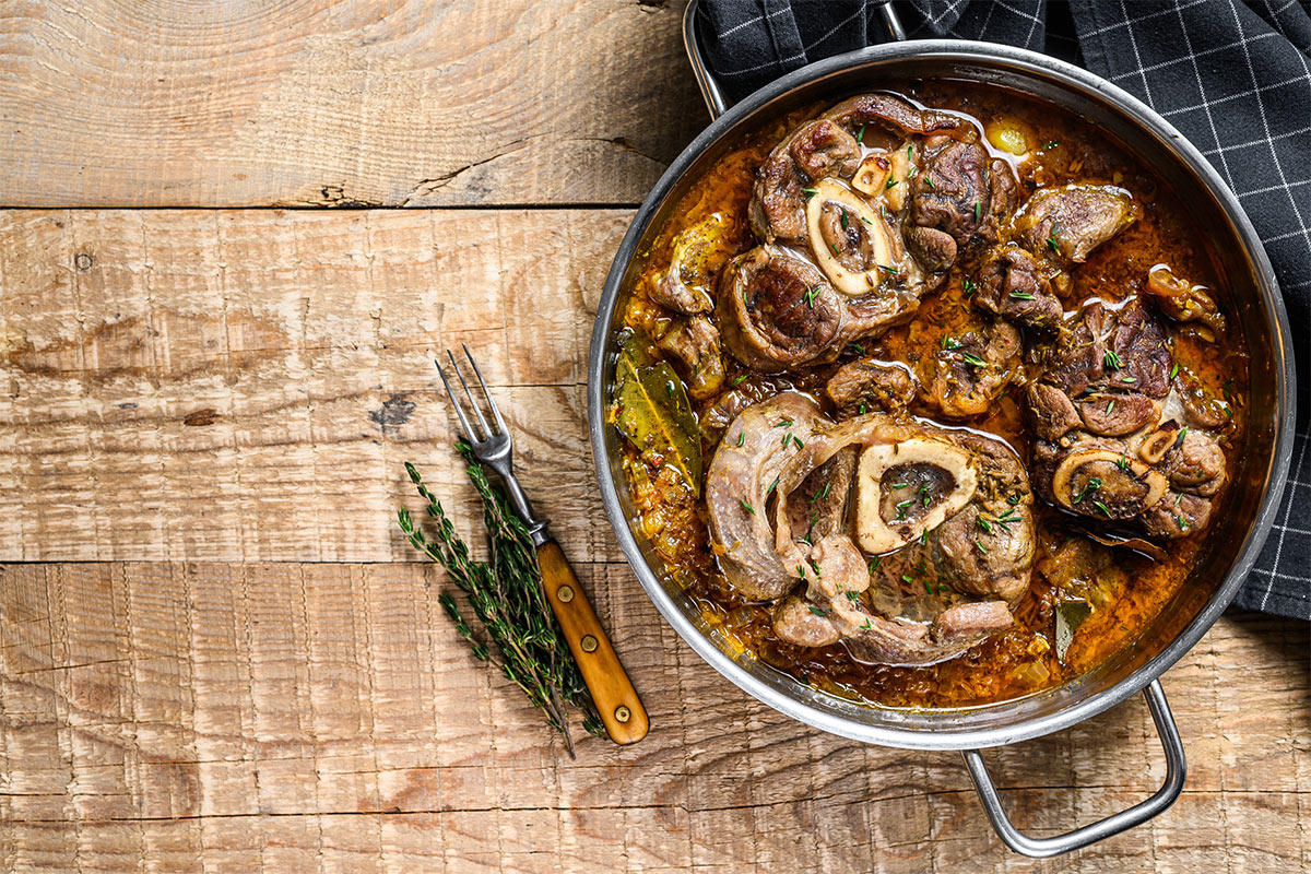 Bird's eye view of a pot with Osso buco on a wooden surface. Next to the pot is a fork, black towel, and some herbs | Girl Meets Food