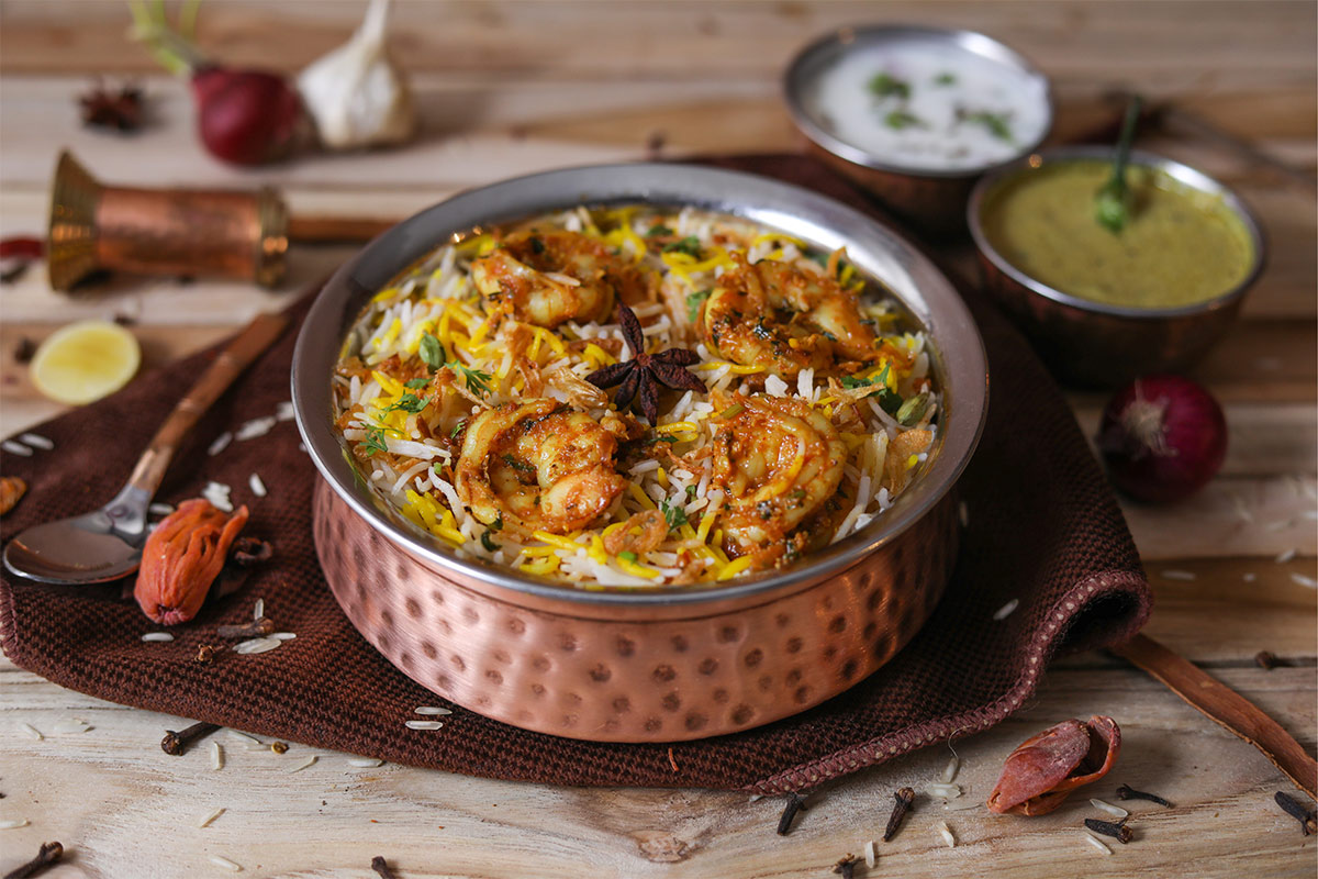 Biryani rice is served in a metal bowl. There are 2 small bowls with sauces next to it | Girl Meets Food