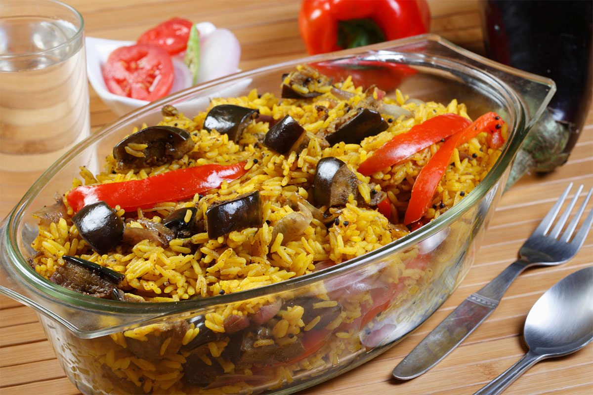 A transparent baking dish full of biryani with vegetables is on a table. There is cutlery, a glass of water and veggies next to it | Girl Meets Food