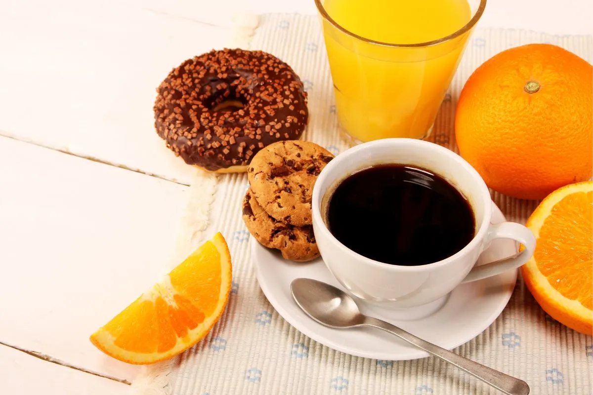 A cup of coffee, a glass of juice, oranges and donut are on a white tablecloth that is on a white wooden surface | Girl Meets Food