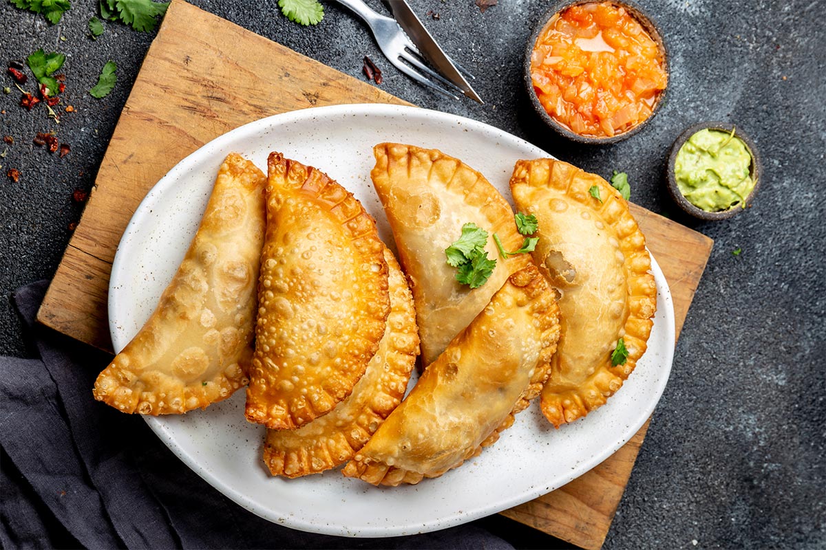 A plate with empanadas is on the cutting board that is on a black surface. There is a fork, knife, and two sauce bowls next to the board  | Girl Meets Food