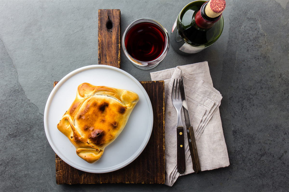 Bird's eye view of a plate with empanadas that is on the cutting board, fork and knife on a linen towel, a glass and bottle of red wine. Everything is on a grey marble surface | Girl Meets Food