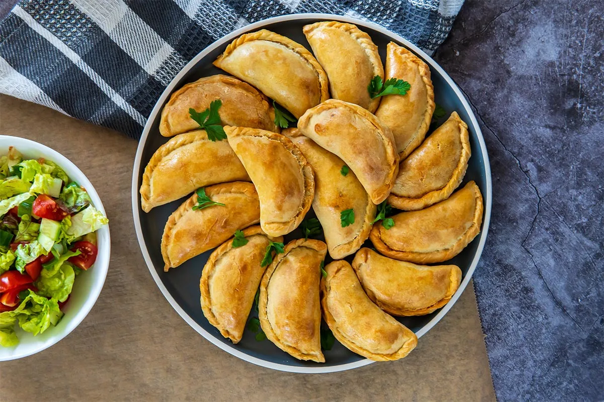 A grey plate with empanadas stands on a perhament paper. There is a bowl of salad and a towel next to it | Girl Meets Food