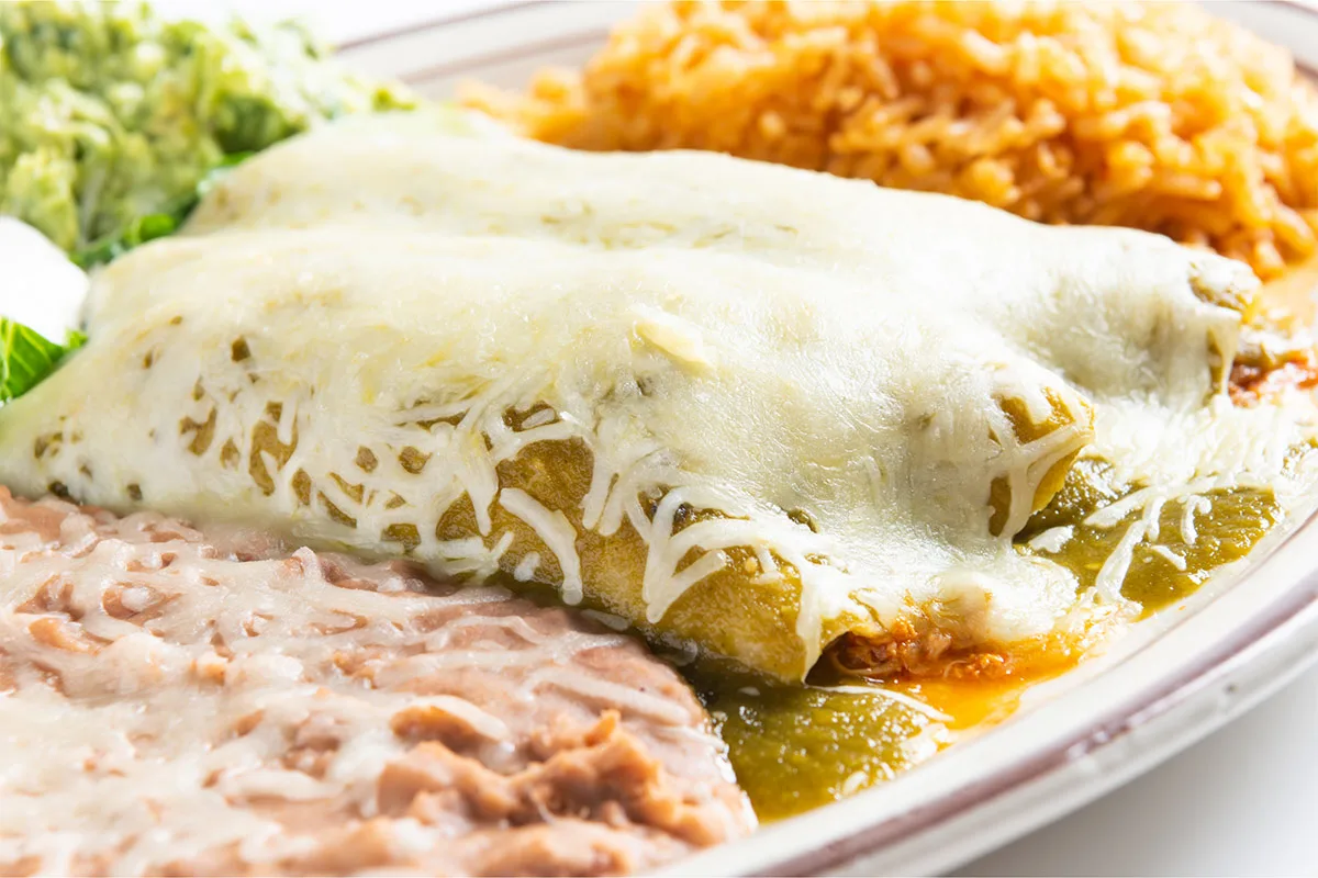 A plate of enchiladas with cheese topping, green sauce and sides is on a table | Girl Meets Food