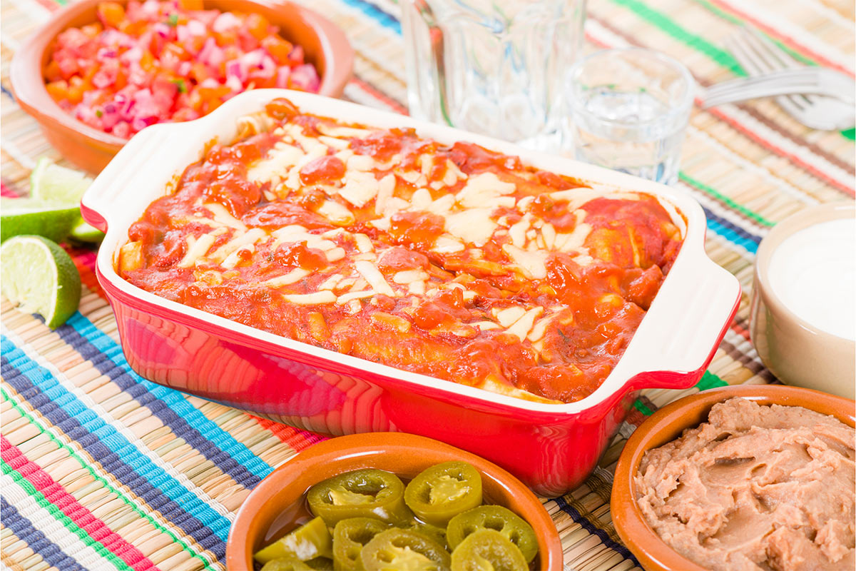 A red baking dish with enchiladas is on a colorful tablecloth. The dish is surrounded with salads and crockery | Girl Meets Food
