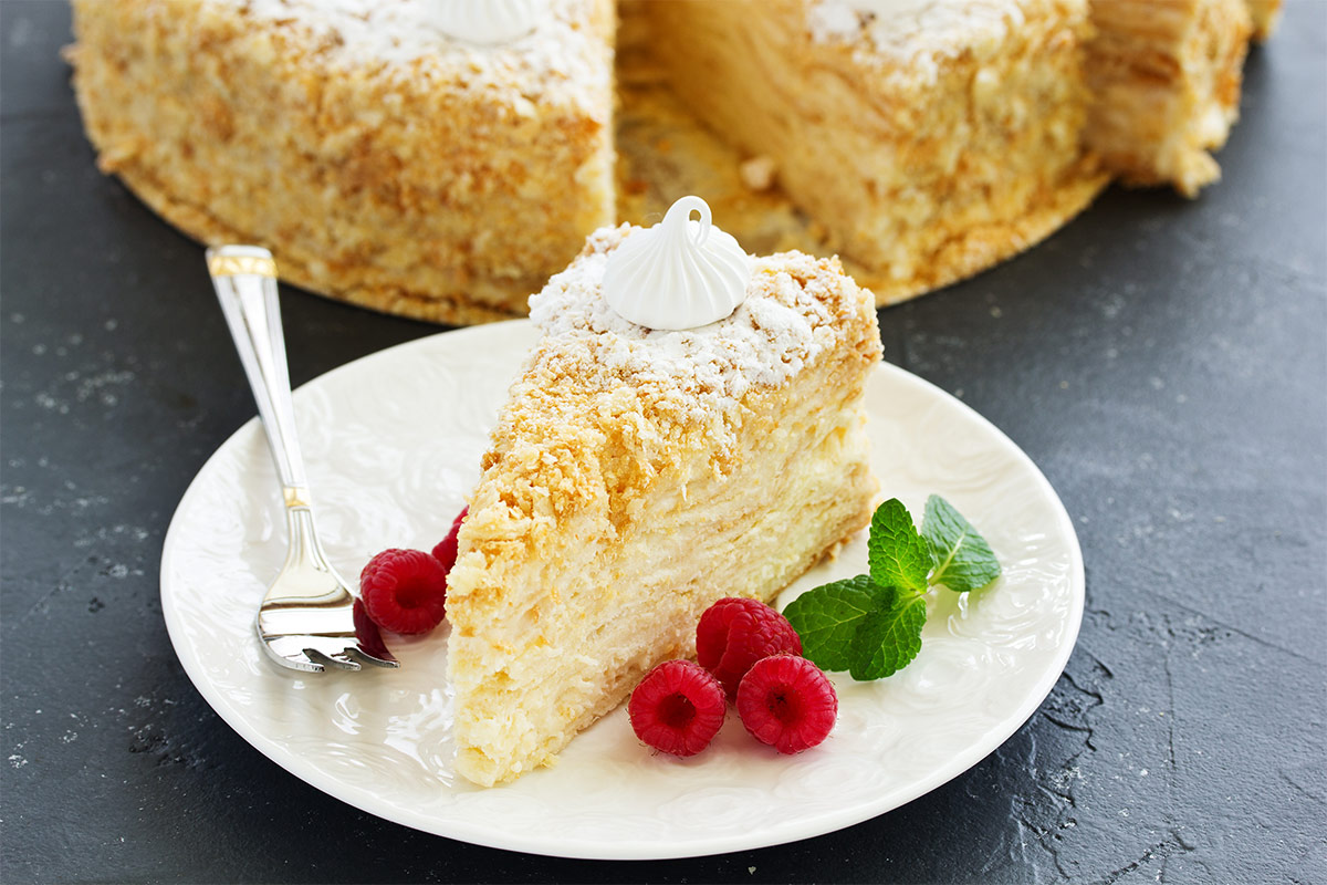 A piece of Napoleon cake is served on a white plate with strawberries mint leaves and a fork. A plate is on a black surface. A Napoleon cake is behind the plate | Girl Meets Food