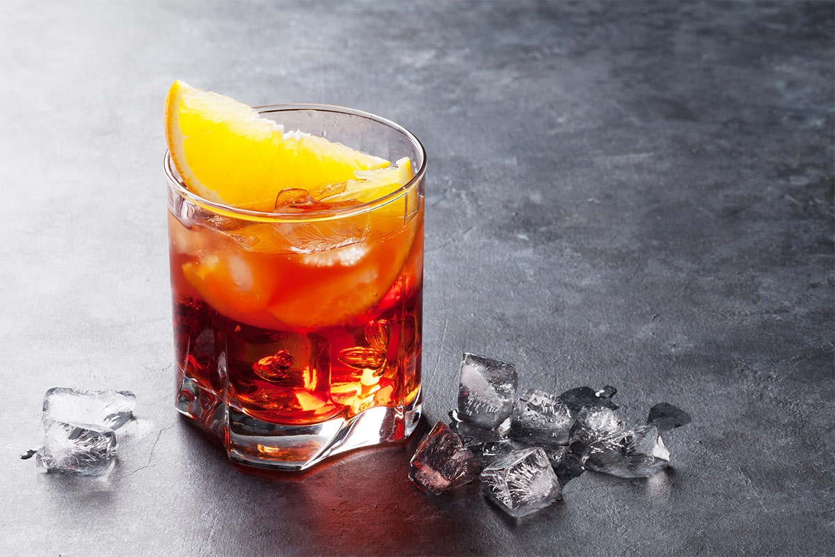 A glass of negroni cocktail is on a grey surface with some ice cubes alongside it | Girl Meets Food