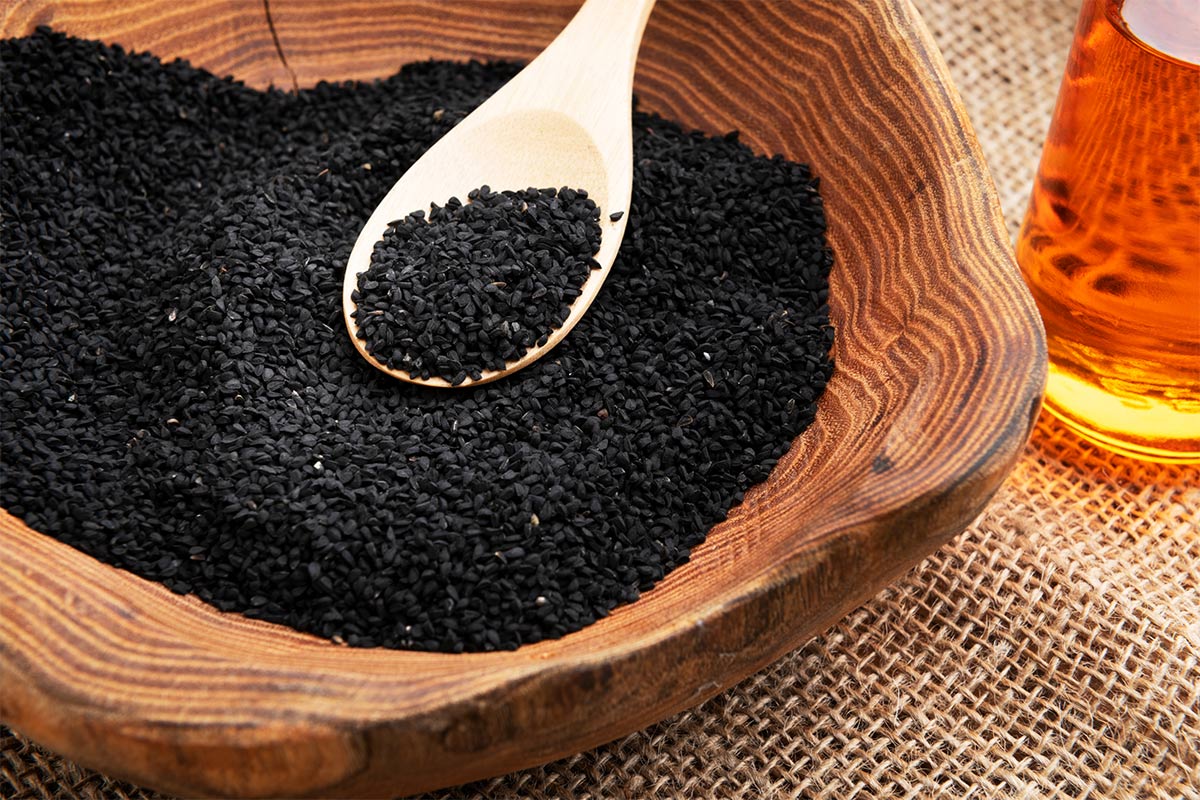 Nigella seeds in a wooden bowl with wooden spoon. The bowl stands on a linen tablecloth. There is a bottle of oil next to the bowl | Girl Meets Food