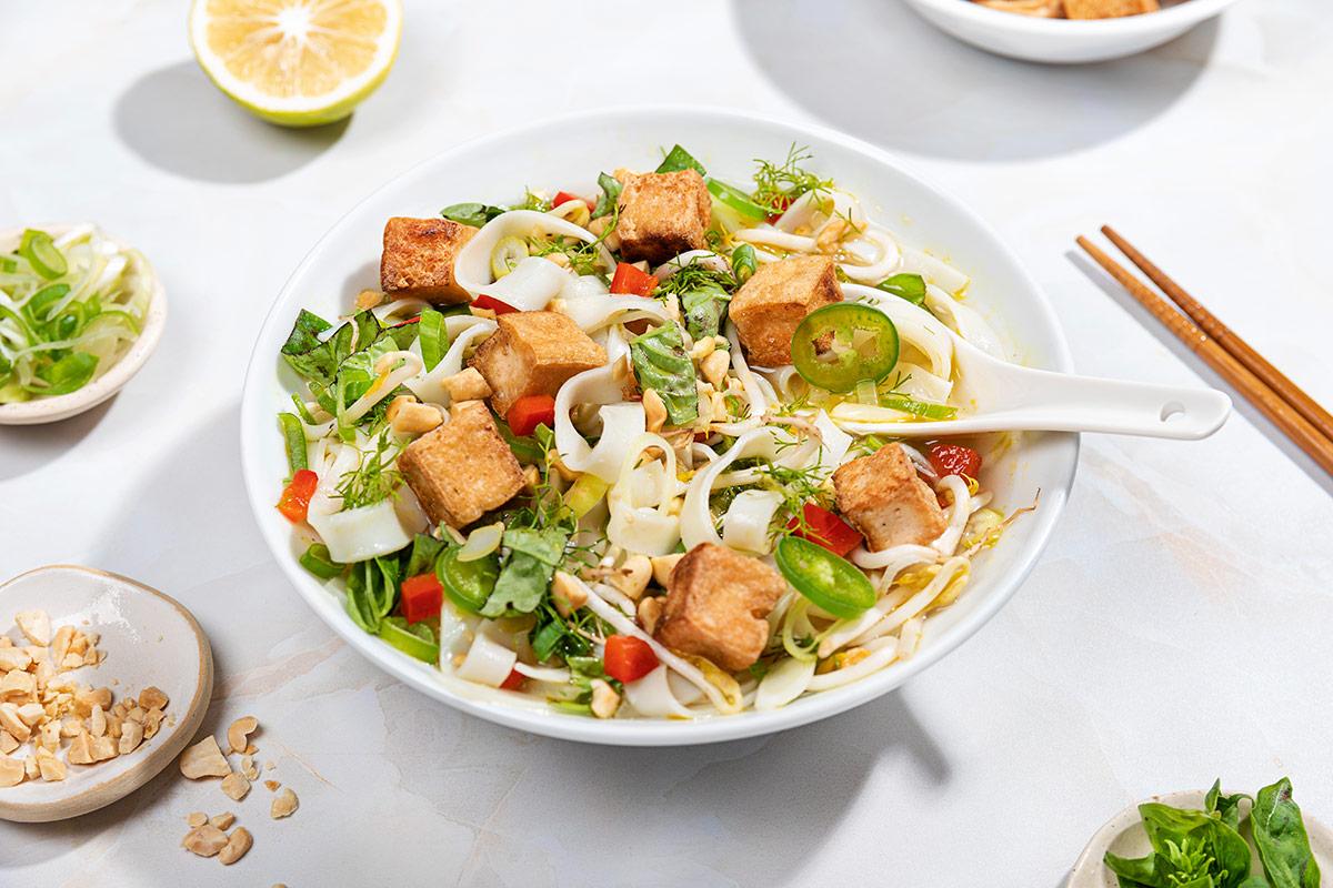 A bowl of vegan noodle soup with a ceramic soup and spoon sunk into it. Small trays of toppings surround it | Girl Meets Food