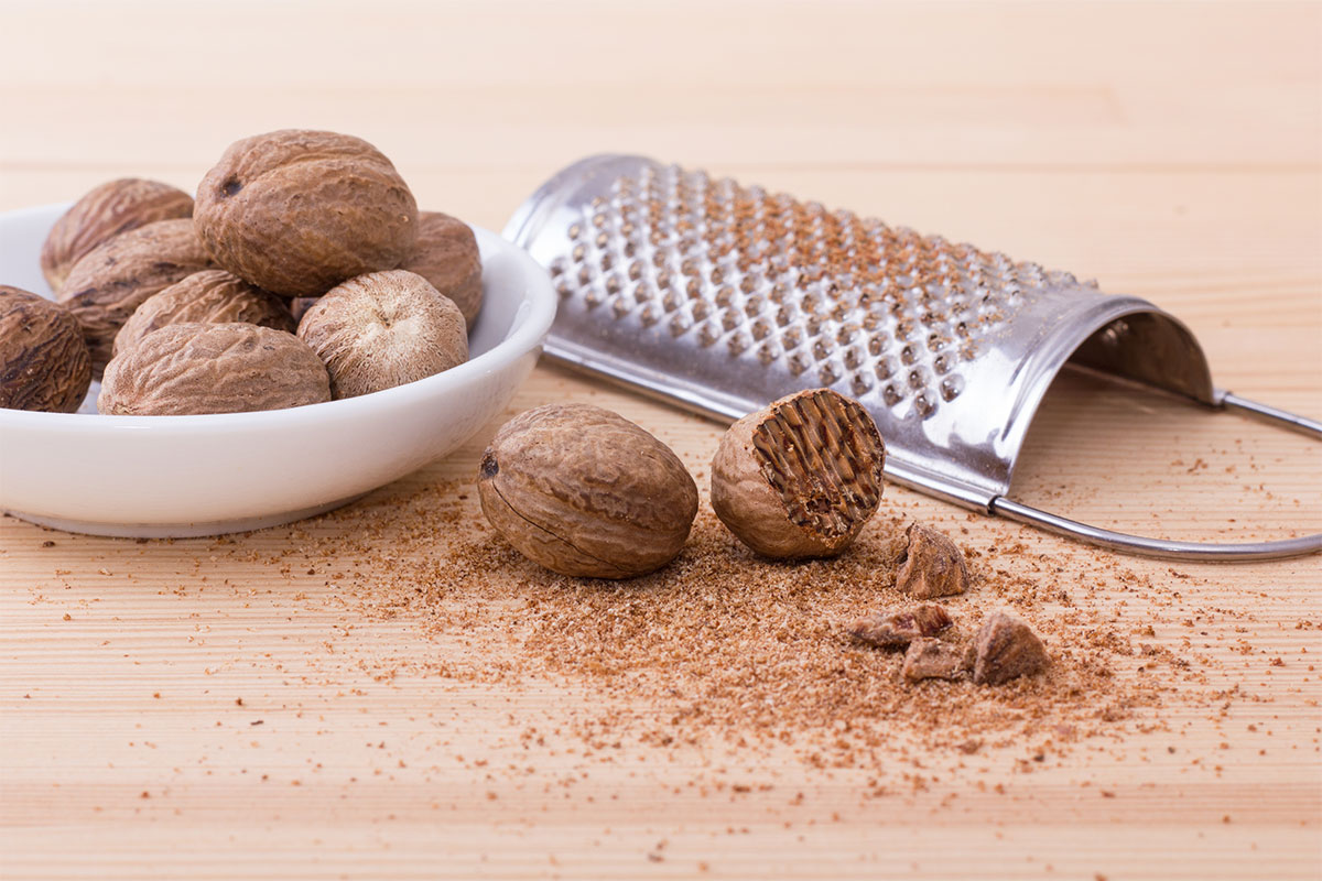 Several nutmegs are in a bowl that is on a wooden surface. Two nutmegs and a grater are next to the bowl | Girl Meets Food