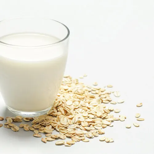 A glass of oat milk is on a white surface. Oats are scattered around the glass | Girl Meets Food