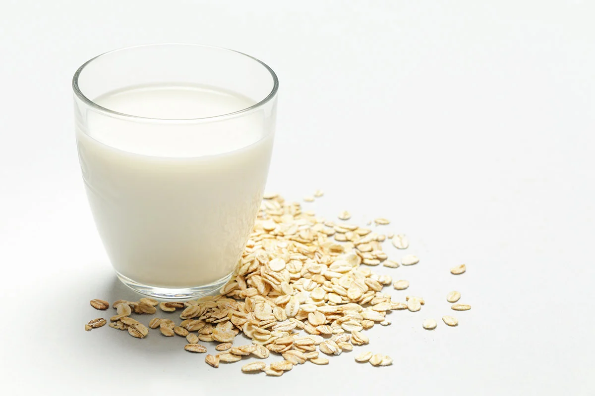 A glass of oat milk is on a white surface. Oats are scattered around the glass | Girl Meets Food