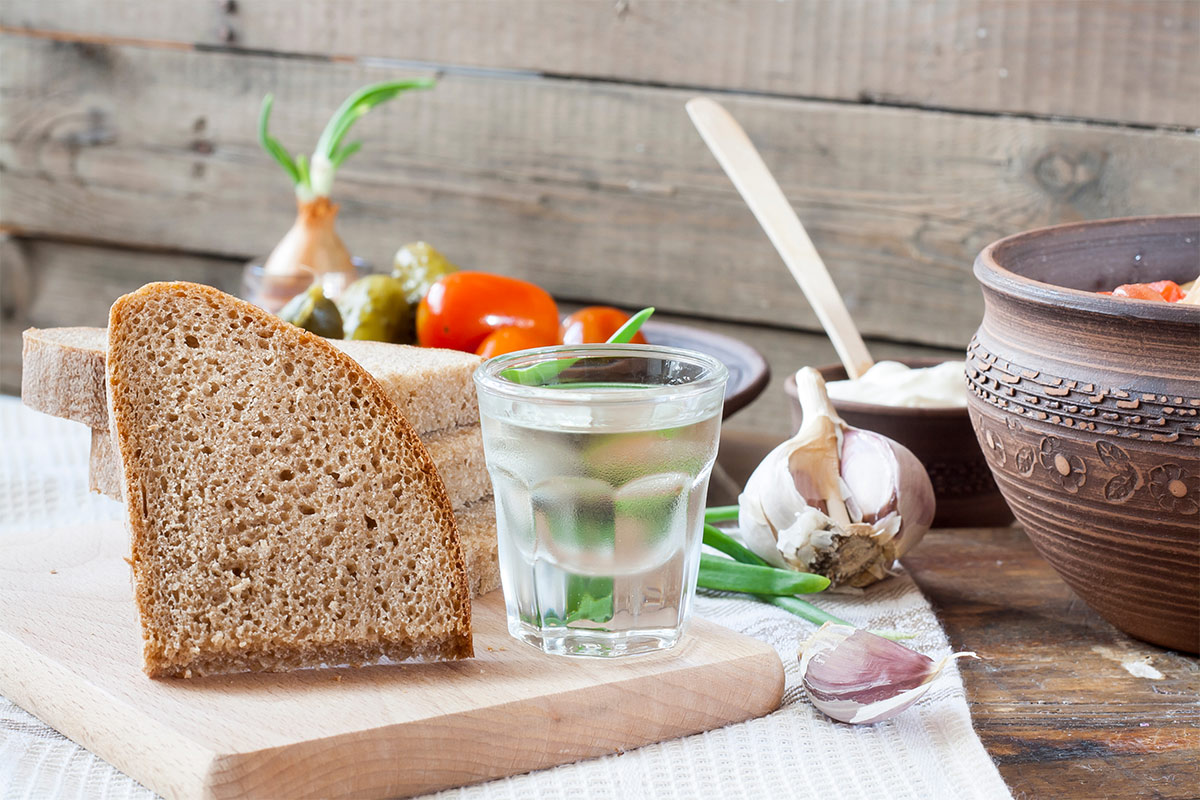 A glass of horilka with sliced bread on the cutting board. There are some dishes on the background | Girl Meets Food