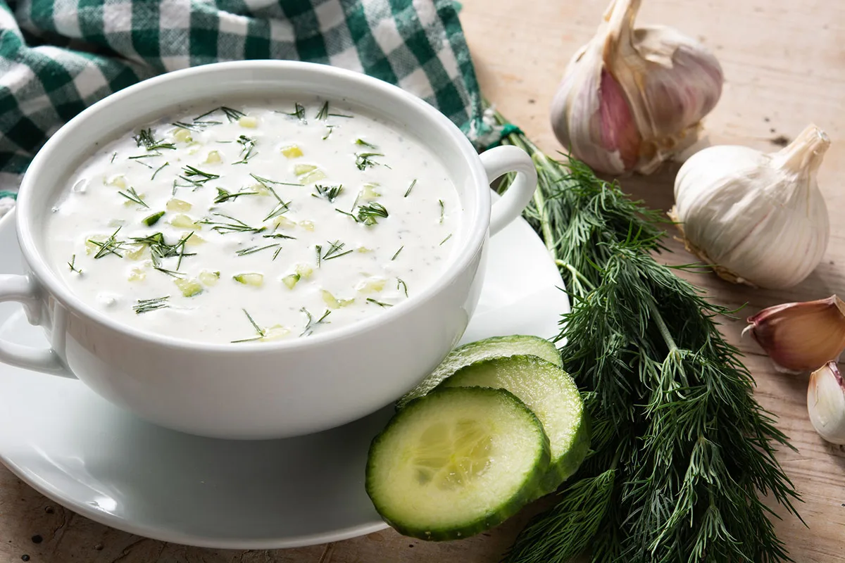 A bowl of Tarator soup surrounded with garlic, dill, cucumber slices and a towel on the table | Girl Meets Food