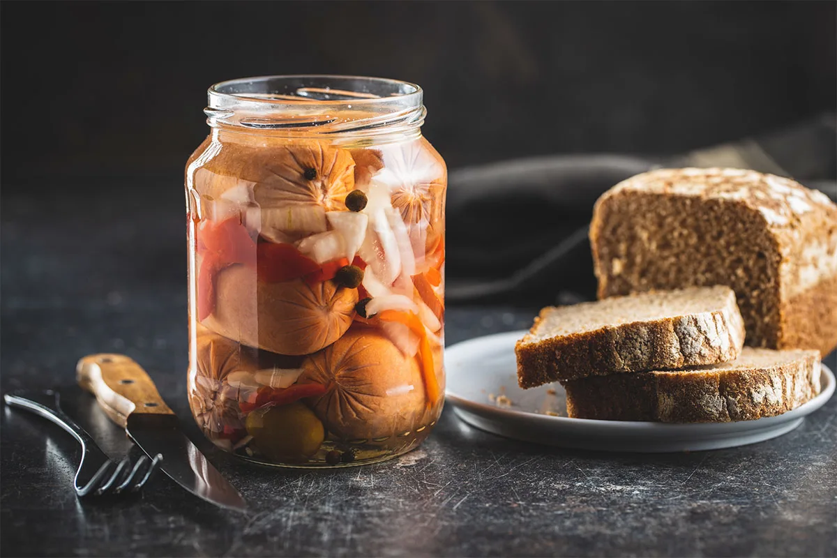 A jar of Utopenec (pickled sausages) on the black surface. There are knife, fork and a plate of sliced bread next to it | Girl Meets Food
